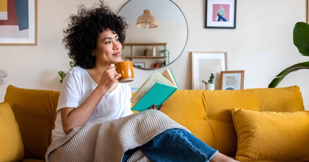 Pensive relaxed African american woman reading a book at home, drinking coffee sitting on the couch. Copy space. Lifestyle concept.