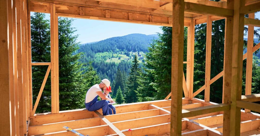 Father with toddler son building wooden frame house near forest. Male builder showing his son the construction plan, wearing helmets and blue overalls on sunny day. Carpentry and family concept.