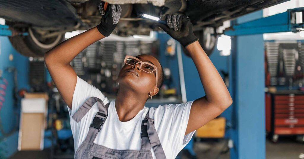 A young African American female mechanic is standing under a car and examining the damage on the broken parts with a serious look on her face.
