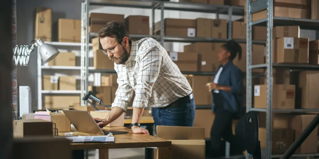 Successful Small Business Owner of a Retail Online Shop Preparing a Small Cardboard Parcel for Postage. Stylish Young Inventory Manager Working on Laptop Computer in Warehouse Facility.